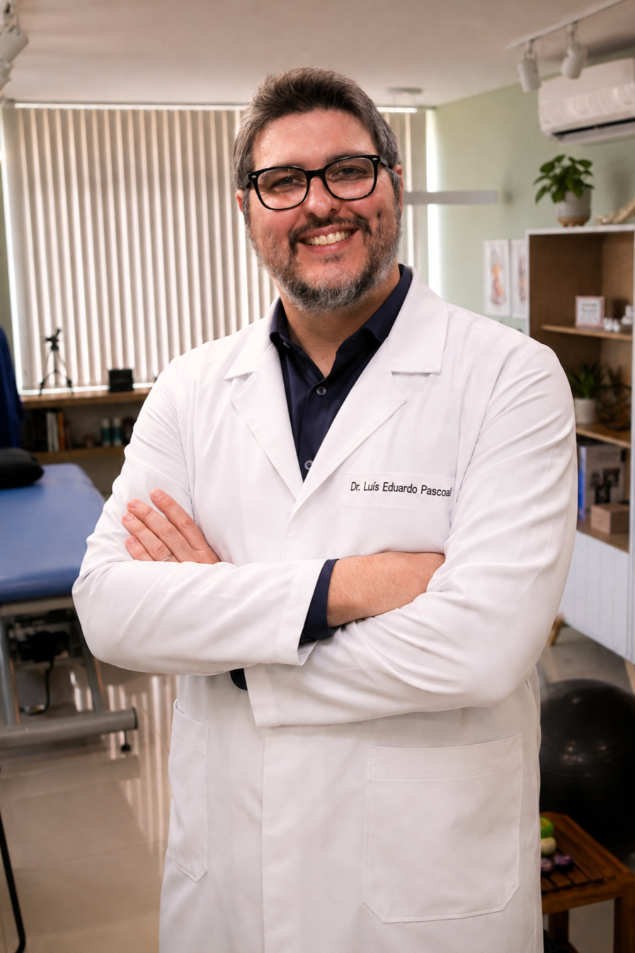 Dr. Luís Eduardo Pascoal, fisioterapeuta da equipe EARFF de osteopatia, homem sorrindo de óculos e barba, vestindo scrub azul-marinho, sentado à mesa em consultório clínico com estante de livros e persiana ao fundo, fotografia realista