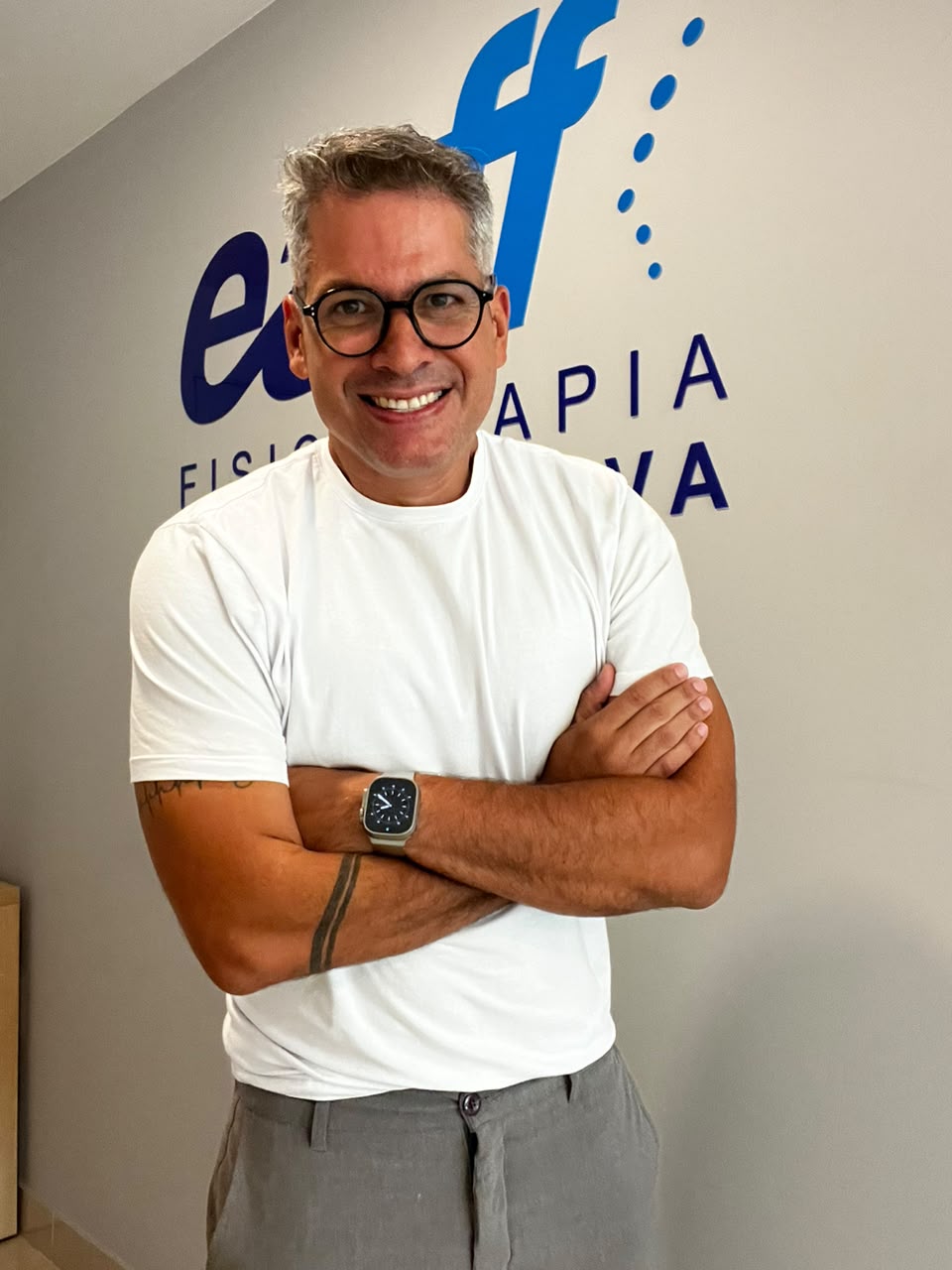 Dr. Fernando Capristano, fisioterapeuta da equipe EARFF, homem sorrindo de óculos de cabelo grisalho, braços cruzados, vestindo camiseta branca, em frente a parede com logo da clínica, fotografia realista