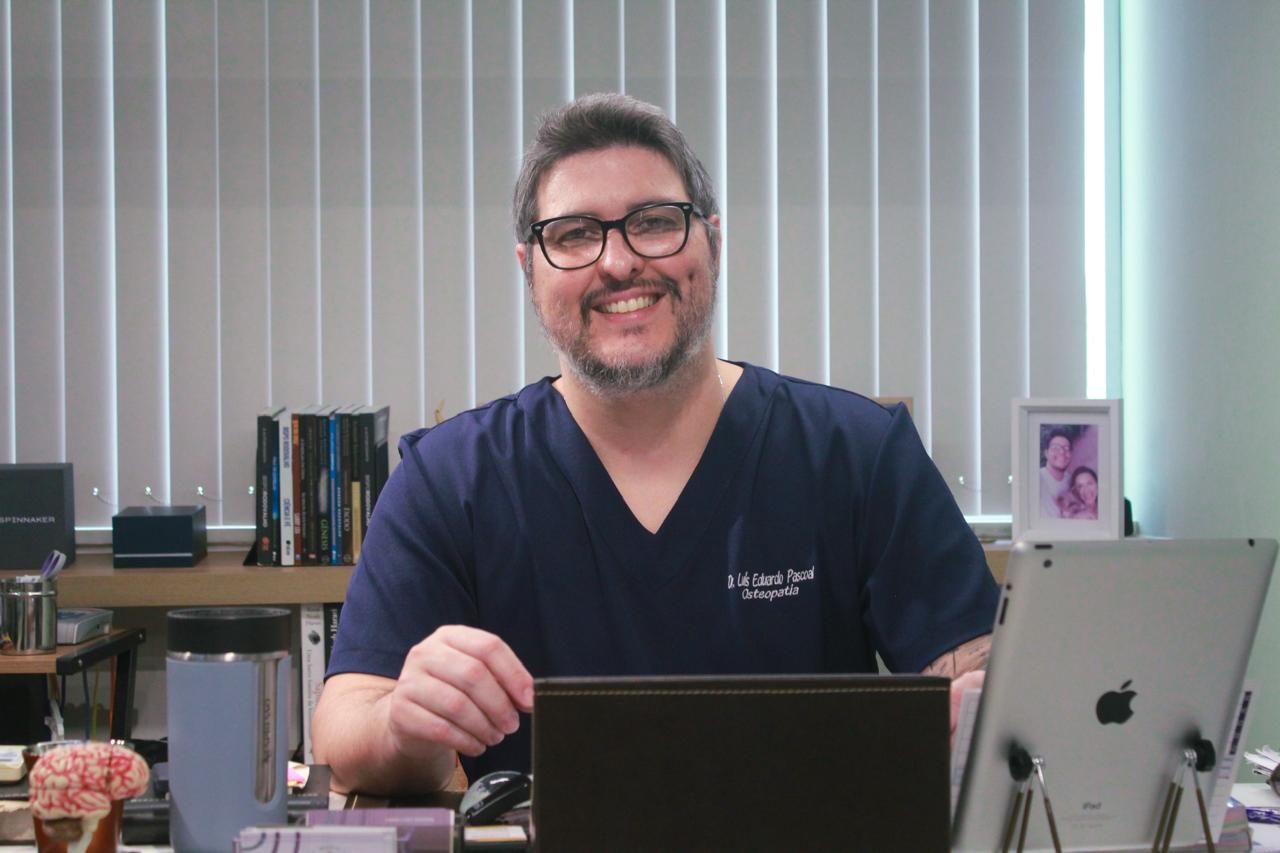 Dr. Luís Eduardo Pascoal, fisioterapeuta da equipe EARFF de osteopatia, homem sorrindo de óculos e barba, vestindo scrub azul-marinho, sentado à mesa em consultório clínico com estante de livros e persiana ao fundo, fotografia realista