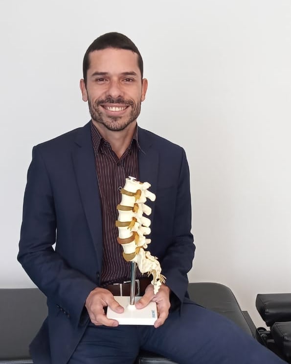 Dr. Angel Mello, fisioterapeuta da equipe EARFF, homem sorrindo de terno azul-marinho segurando um modelo anatômico de coluna vertebral, sentado em ambiente clínico claro, fotografia realista